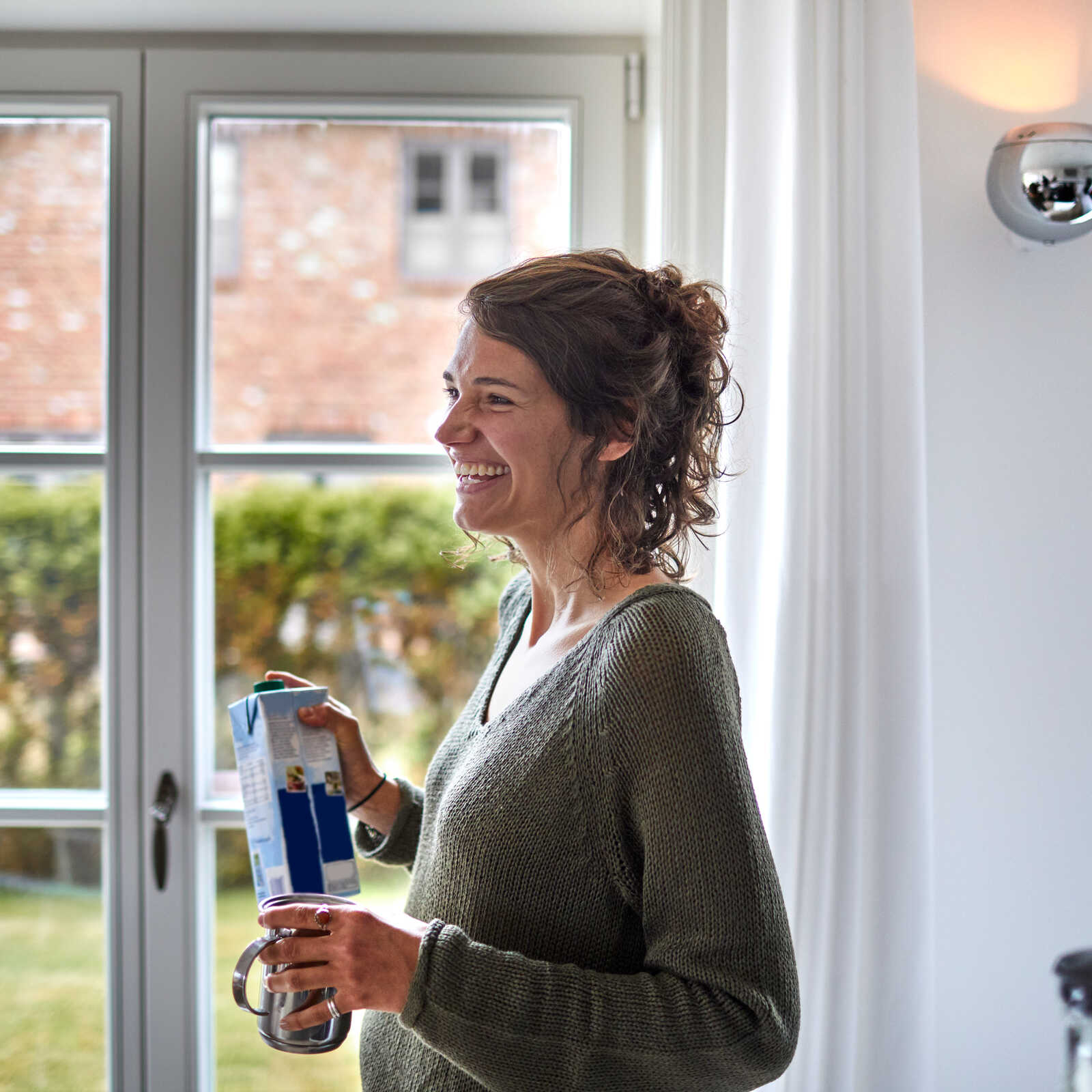 Kvinde smiler ved vinduet med en mælkekarton og krus i hænderne, omgivelserne er lyse og hjemlige med udsigt til haven.