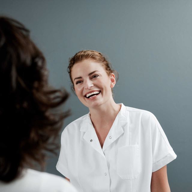 En smilende kvinde iført en hvid skjorte ser glad ud, mens hun taler med en person uden for billedet.