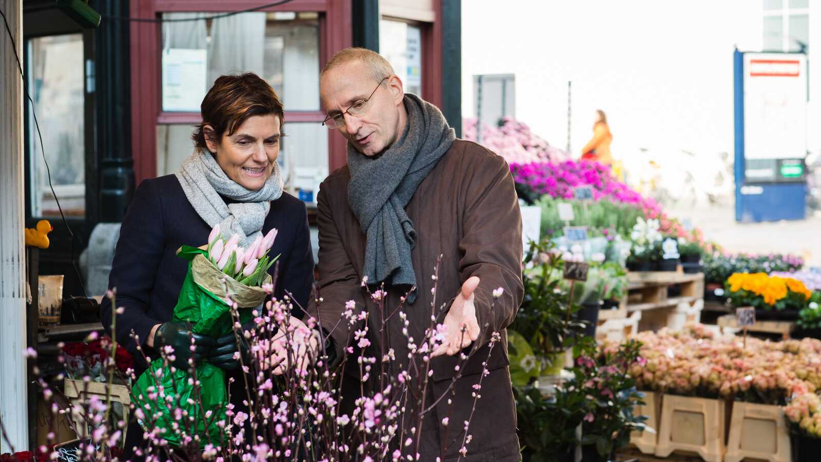 Par står ved en blomsterbod og kigger på blomstergrene. Kvinden holder en buket tulipaner, og manden peger på pink blomster.