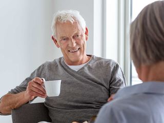 Ældre mand i grå sweater smiler, mens han holder en kop kaffe. Sidder ved et bord med frugtskål og taler med en anden person.