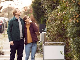 Ungt par står ude og kigger på et hus i efteråret, mens de holder i hånd og ser smilende ud.