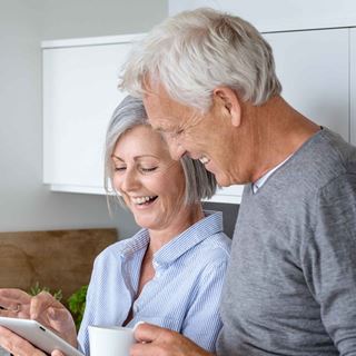 Parret smiler sammen, mens de kigger på en tablet i køkkenet. Kvinden holder en kop, og omgivelserne er lyse og moderne med en hyggelig atmosfære.