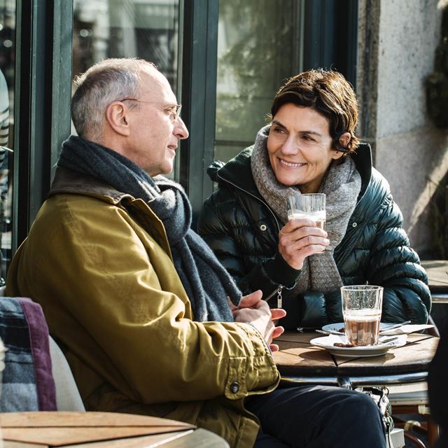 To personer sidder udendørs på en café og nyder kaffe sammen. De er klædt i vinterjakker og halstørklæder, mens de smiler til hinanden.