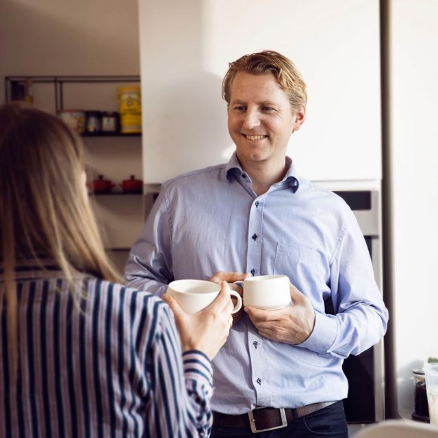 Mand og kvinde står i et køkken og holder hver deres kaffekop. De smiler og taler sammen i et afslappet kontormiljø.