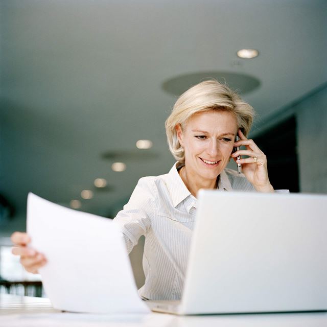 Kvinde arbejder ved en computer, taler i telefon og holder papirer. Moderne kontormiljø med lysloft og minimalistisk indretning.