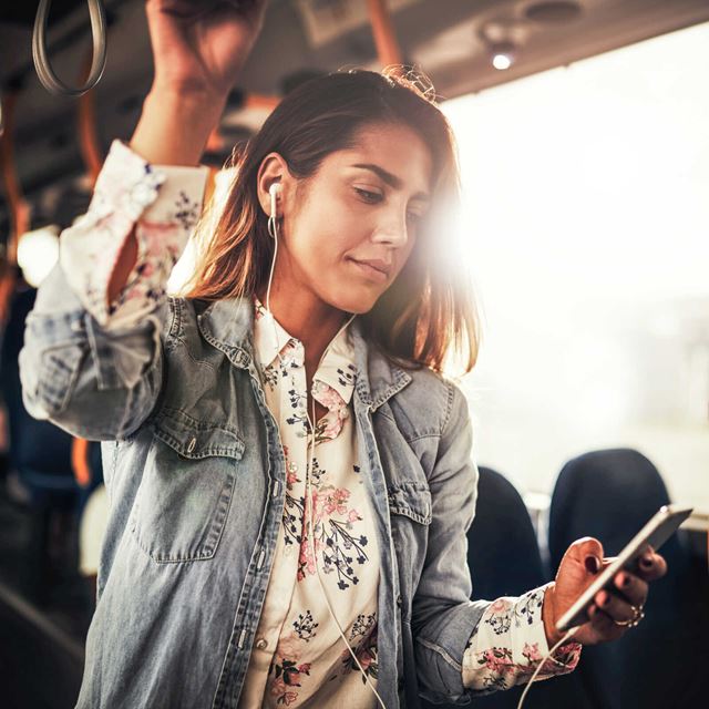 Kvinde i bus med høretelefoner, holder fast i stroppen og bruger mobiltelefonen mens morgensolen skinner gennem vinduet.