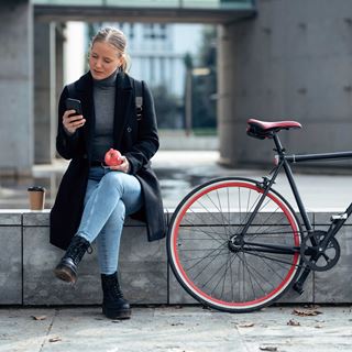 En kvinde sidder ved siden af sin cykel, spiser et æble og kigger på sin telefon med en kop kaffe ved siden af sig.