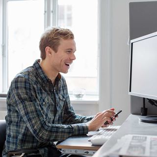 Begejstret ung mand smiler, mens han arbejder ved sin computer og holder sin telefon i hånden.