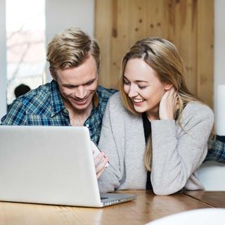 Et ungt par sidder ved et bord, ser på en laptop og en telefon og smiler.