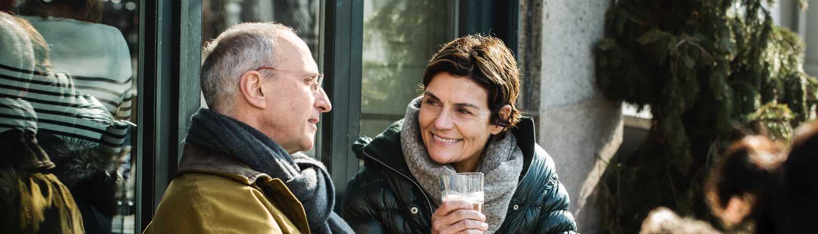 To personer sidder udendørs på en café og nyder kaffe sammen. De er klædt i vinterjakker og halstørklæder, mens de smiler til hinanden.