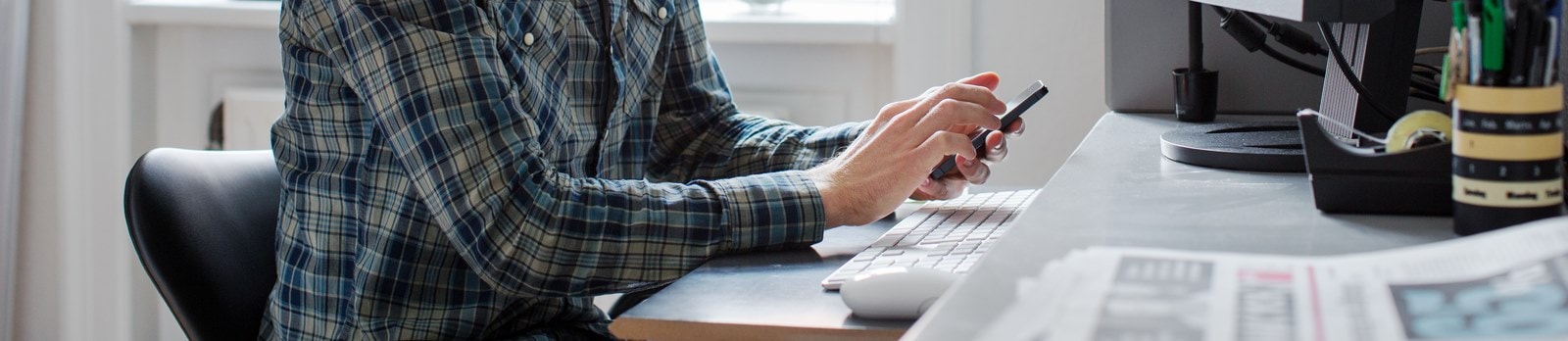 Begejstret ung mand smiler, mens han arbejder ved sin computer og holder sin telefon i hånden.